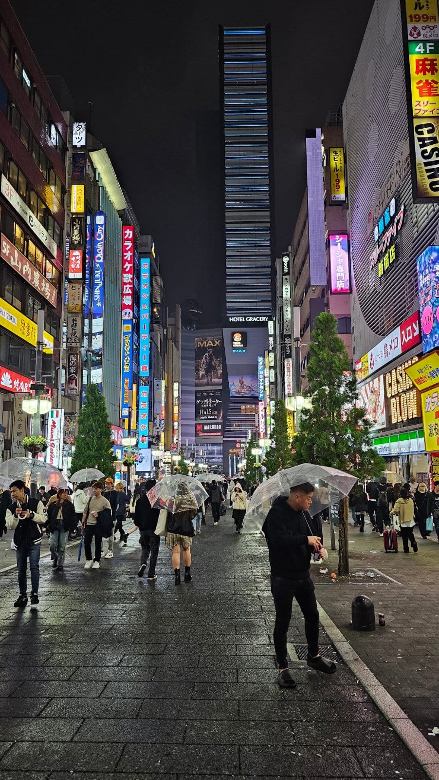 Kabukicho at night, with illuminated signs, Hotel Gracery and pedestrians carrying umbrellas on a wet street
