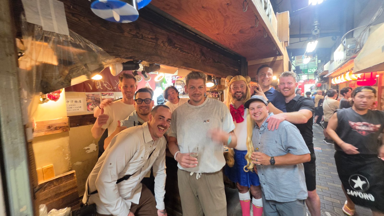 A group smiling together with drinks outside a warmly lit bar in a narrow alley