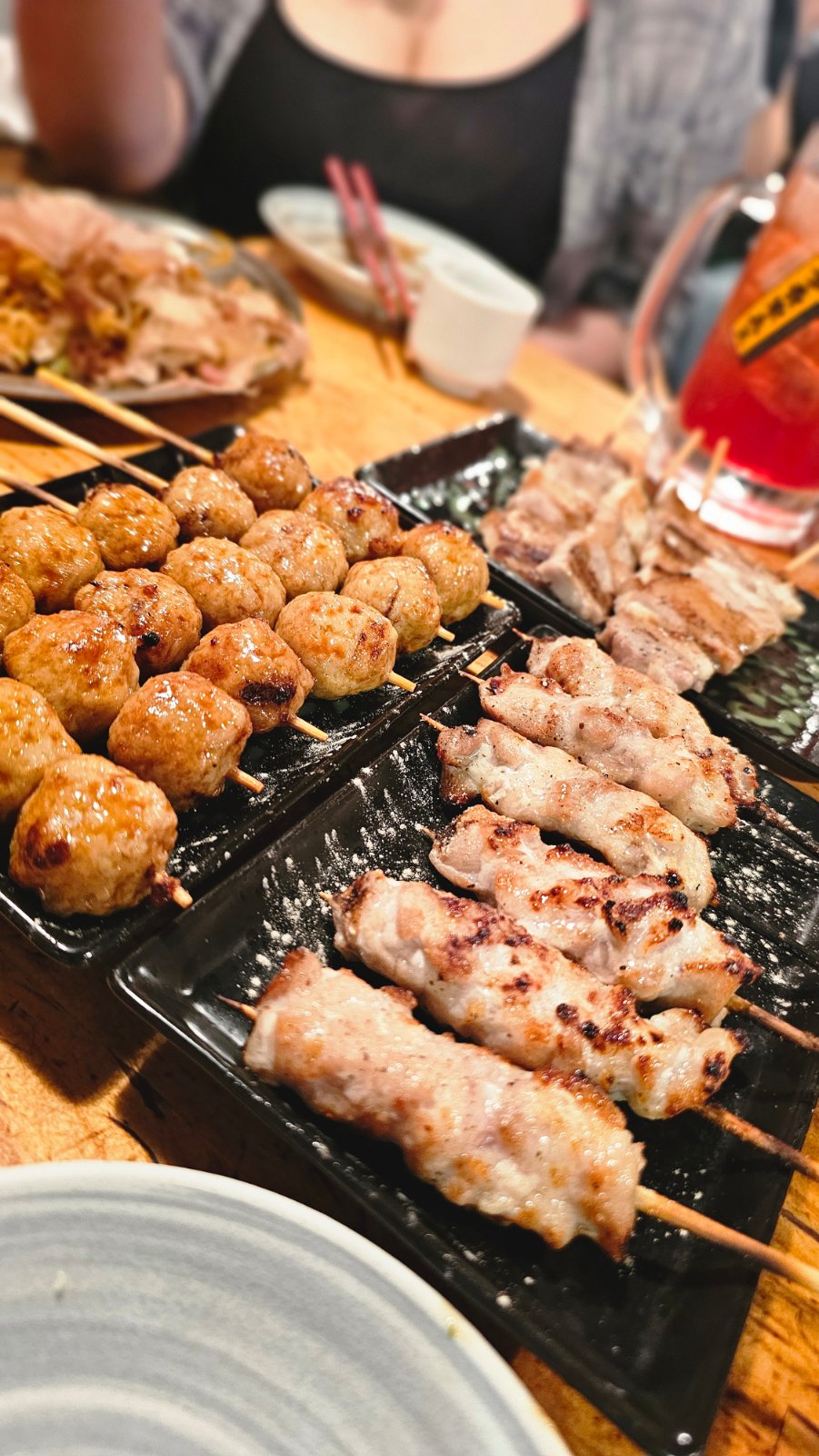 Grilled skewers and glazed meatball skewers on black plates, with a drink on the dinner table