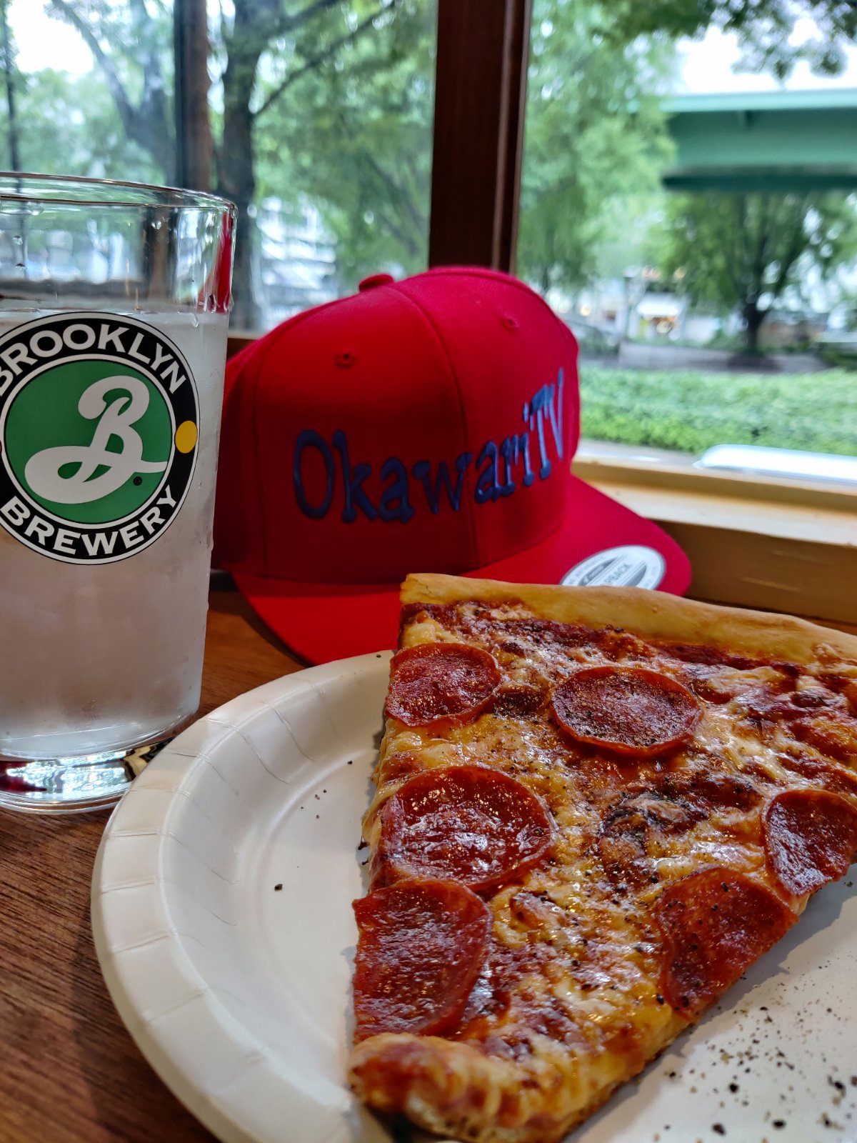 Pepperoni pizza beside a drink and a red Okawari cap by a window