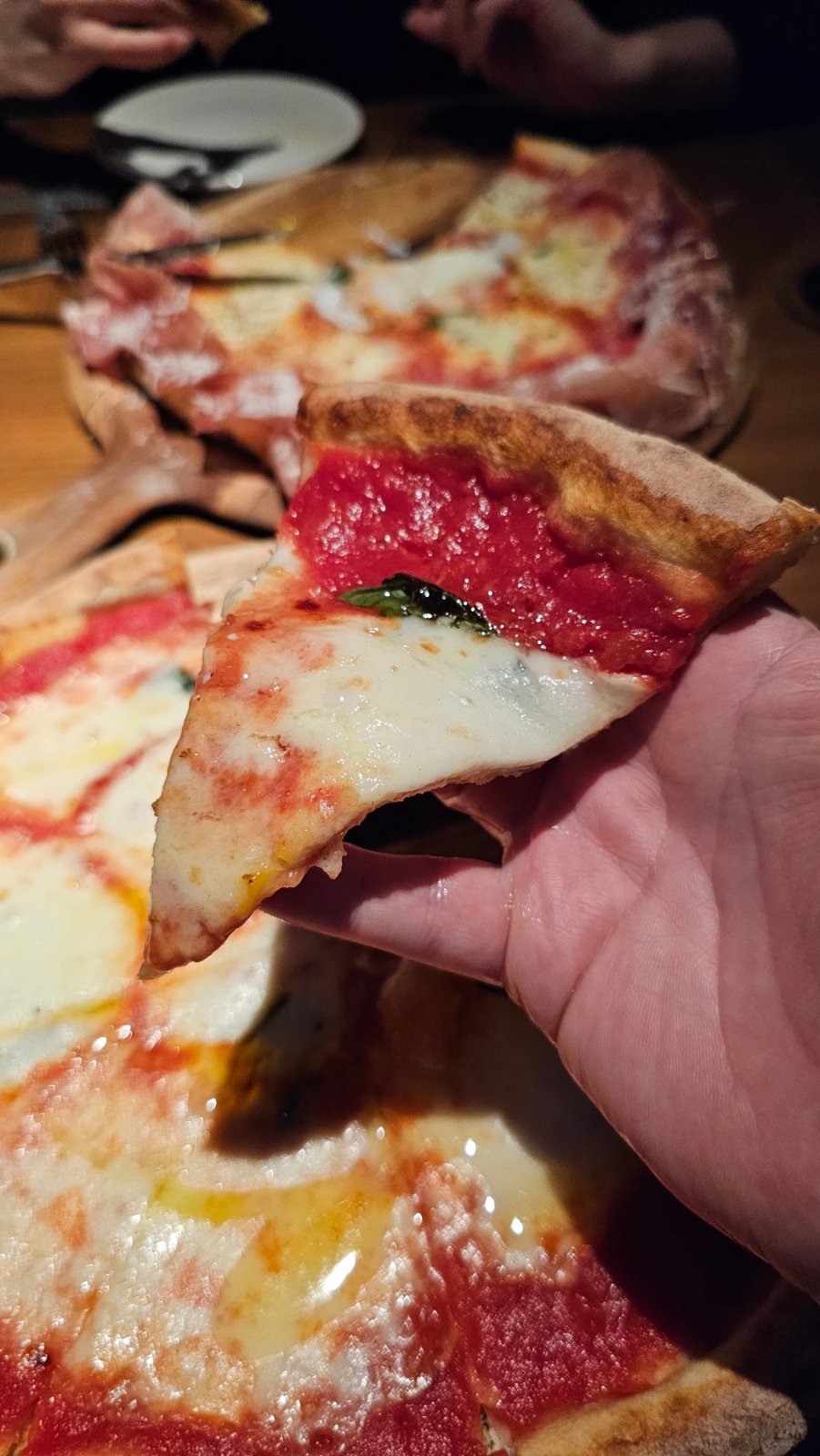 A hand holding a slice of tomato, cheese and basil pizza, with pizzas on the table behind it