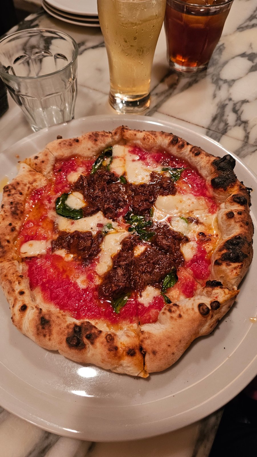 Pizza with a blistered crust, tomato, cheese and basil on a white plate, with drinks behind it