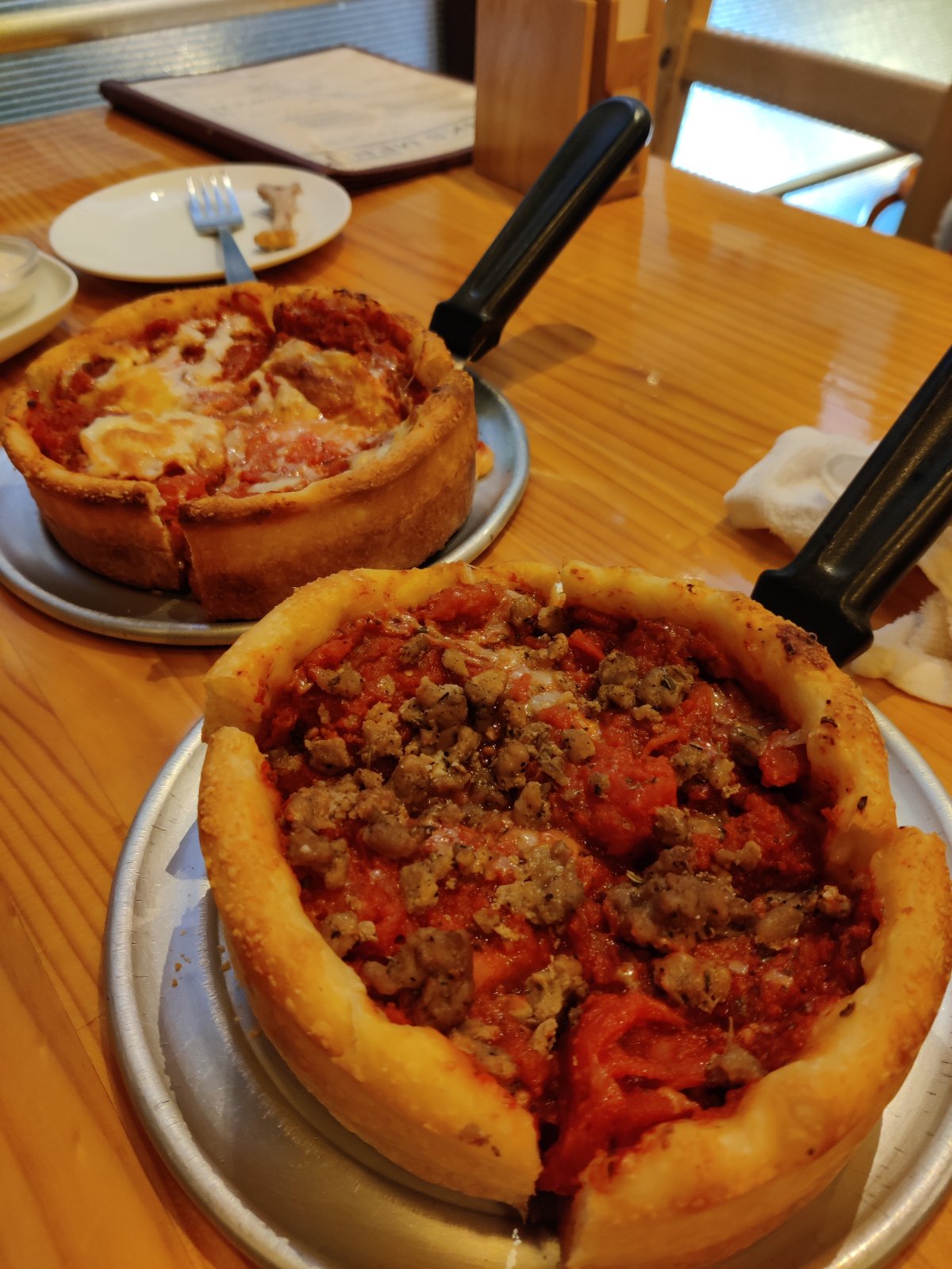 Two deep-dish pizzas on metal serving trays on a wooden table