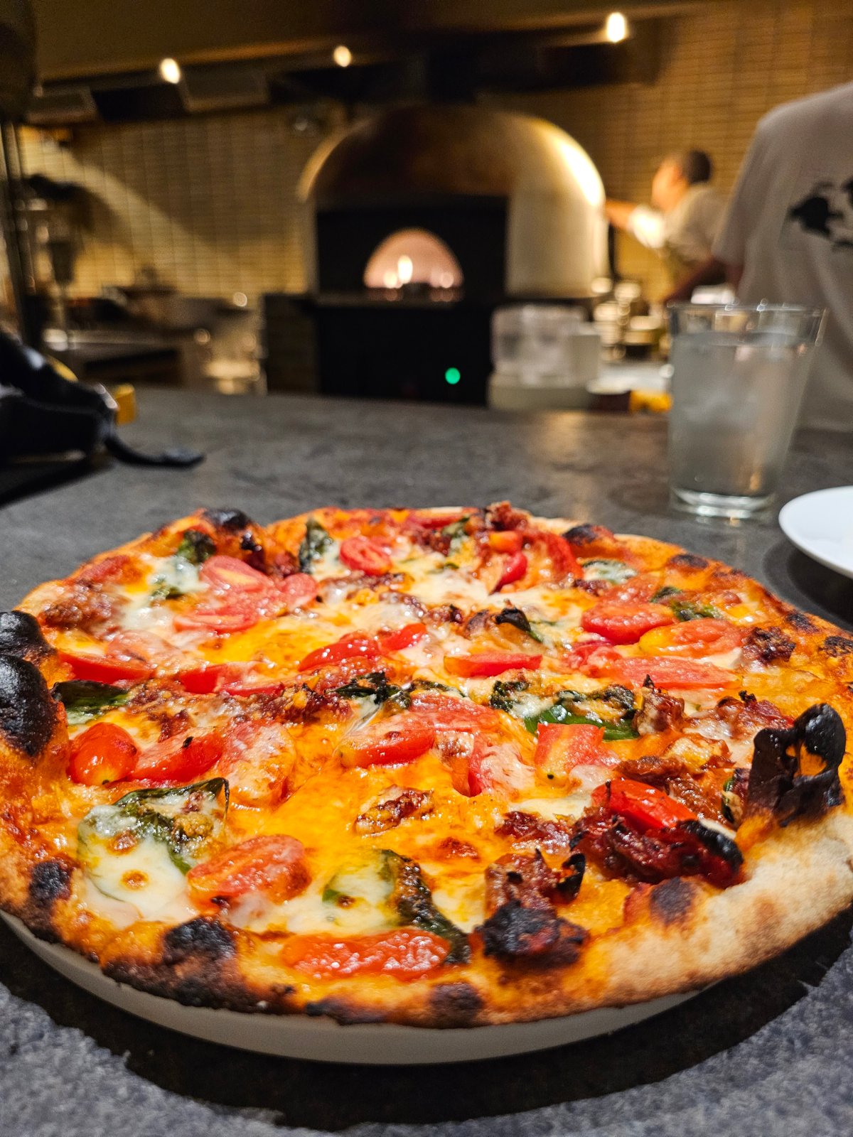 Pizza topped with tomatoes, cheese and basil, with a pizza oven in the background